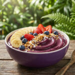 Un bol d'açaï violet, richement garni de tranches de banane, fraises, myrtilles et granola, sur une table en bois devant une végétation luxuriante.