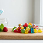 Une brosse à dents, des bonbons colorés, une framboise et de la menthe sur une planche en bois, symbolisant le rapport entre plaisir et santé dentaire.