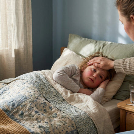 Une mère réconforte son enfant fiévreux au lit, vérifiant sa température. Un verre d'eau et un thermomètre sont sur la table de chevet.