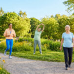 Trois femmes de générations différentes s'adonnent à des activités physiques (course, étirement, marche) dans un parc verdoyant et fleuri sous le soleil.