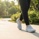 Pieds d'une personne marchant sur un chemin extérieur. Elle porte des baskets grises et un pantalon noir. Lumière naturelle sur la végétation.