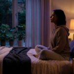 Femme méditant sur son lit dans une chambre sombre avec diffuseur. Ambiance propice à la relaxation et au sommeil.