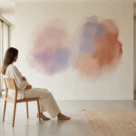 Femme enceinte assise pensivement dans un intérieur lumineux et minimaliste, les mains sur le ventre. Deux taches d'aquarelle colorent le mur.