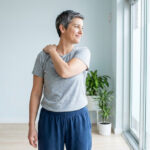 Femme aux cheveux courts gris massant son épaule gauche, cherchant à soulager une douleur, dans une pièce lumineuse près d'une fenêtre.